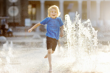 Little boy plays in the square between the water jets in the dry fountain at sunny summer day....