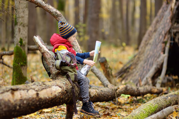 Little boy scout is orienteering in forest. Child is sitting on fallen tree and looking on map on background of teepee hut. Adventure, scouting and hiking tourism for kids.