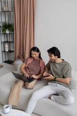 Happy young employees with mobile gadgets discussing online information on laptop screen while sitting on sofa and working from home