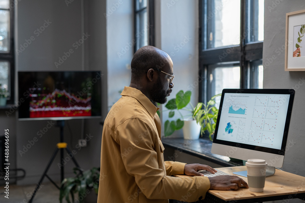 Aufkleber Young modern economist in casual clothes working with financial data in front of computer while sitting by desk in office