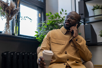 Contemporary young urban office worker with drink talking on smartphone while sitting in armchair...