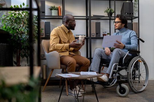 African-American businessman with drink communicating with his Caucasian colleague in wheelchair at working meeting in urban office