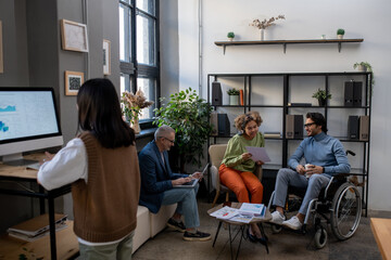 Group of intercultural coworkers in smart casualwear working in urban office while two of them discussing financial data in document