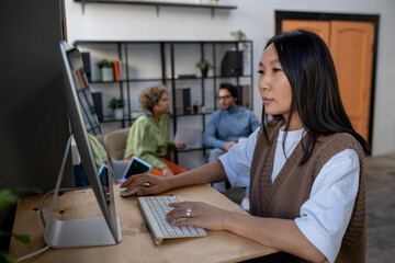 Young Asian businesswoman in casual clothes working in front of computer monitor on background of two intercultural coworkers