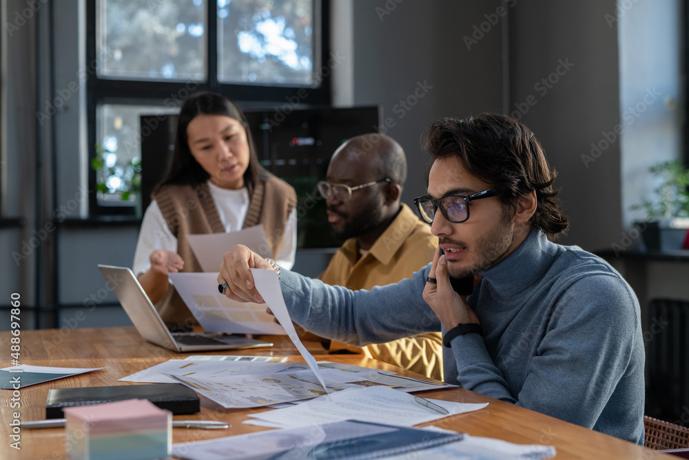 Poster young busy economist with mobile phone by ear looking through data in financial document against two