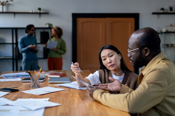 Serious young Asian economist pointing at paper during discussion of data with African-American male colleague at working meeting
