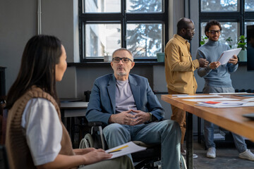 Mature businessman in wheelchair listening to young female colleague at working meeting while sitting against two intercultural coworkers