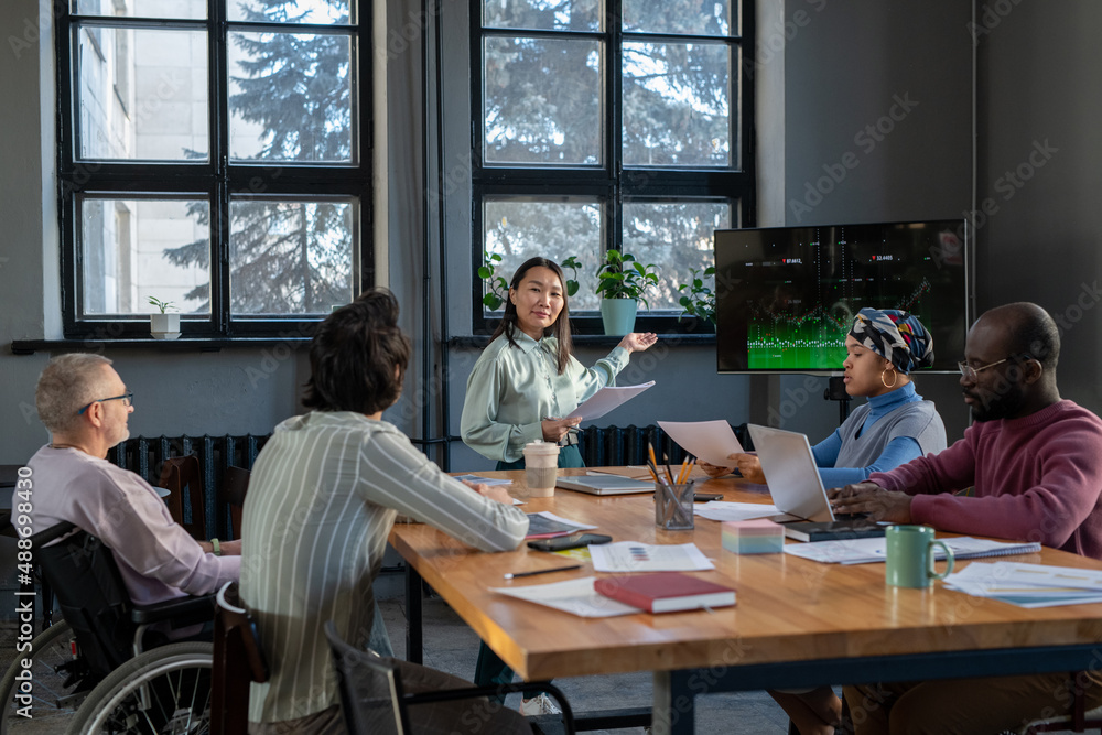 Poster young asian businesswoman with documents pointing at interactive screen with graph while explaining 