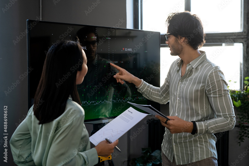 Poster young confident male economist pointing at graph on interactive screen during presentation of financ