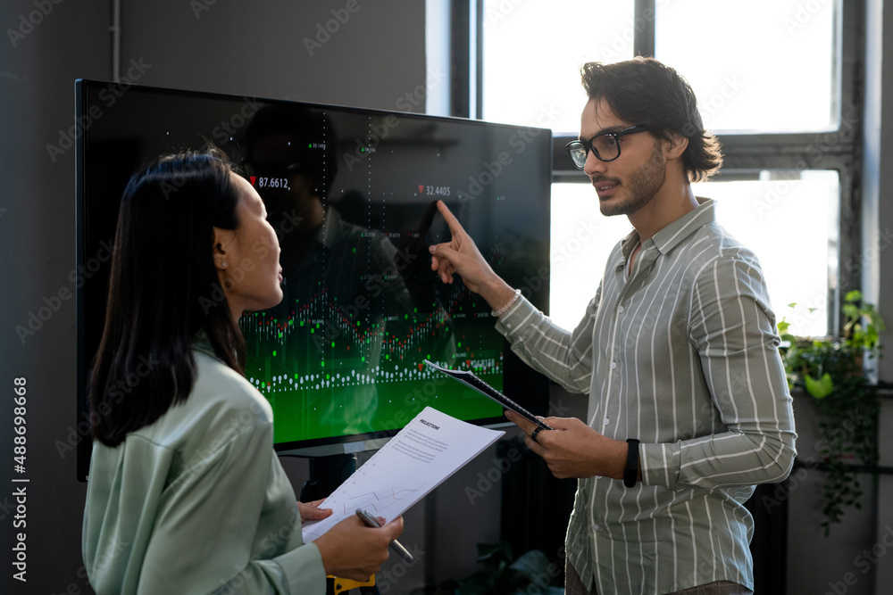 Poster Young confident analyst pointing at digital financial data on interactive screen during presentation and explanation to colleague