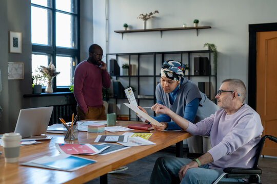 Mature businessman in wheelchair pointing at document held by young biracial female colleague during discussion of financial data