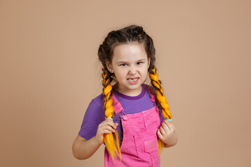 Portrait of angry little, female holding yellow kanekalon braids with vicious eyes looking at...