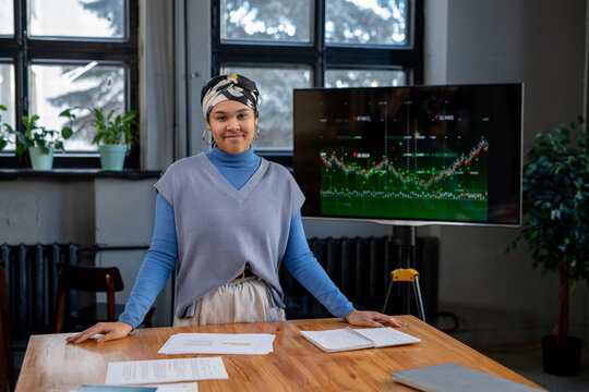 Young contemporary business coach standing by table during presentation of financial data and looking at camera in boardroom - Powered by Adobe