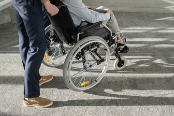 Young elegant businessman standing behind wheelchair with female colleague while helping her move inside modern office center