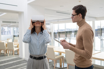 Young businessman with tablet standing by female colleague in vr goggle and entering data in digital database during work over project