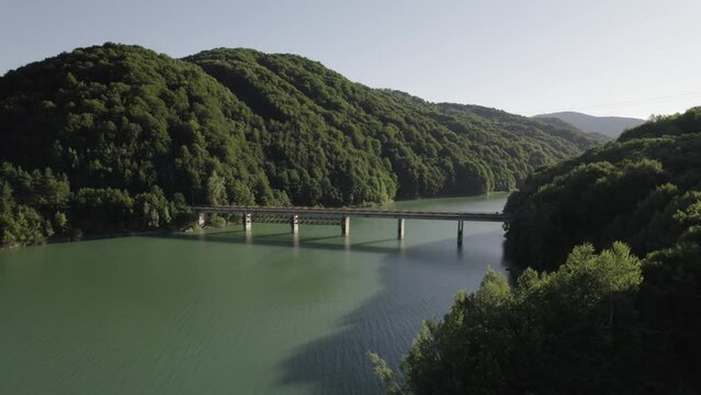 Transalpina highway, steel and concrete beam bridge over Oasa Lake, Romania. Aerial drone shot on a clear day, drone camera aproaches the bridge. Huge lake surrounded by big green mountains.