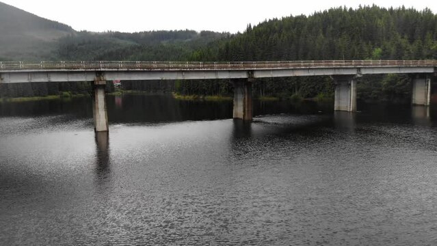 drone shot flying under steel and concrete beam bridge over the Oasa Lake, Romania, part of the transalpine highway. drone goes below the bridge. Huge lake surrounded by pure green pine forest.