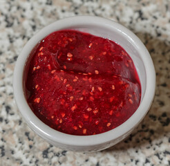 Raspberry jam in a white small bowl on a stone table top view.
