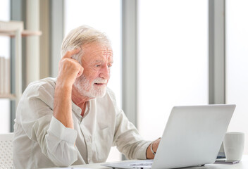 Fototapeta premium Portrait of Worried senior man using laptop and smartphone at home, Mature man in living room with laptop browsing internet on modern computer gadget