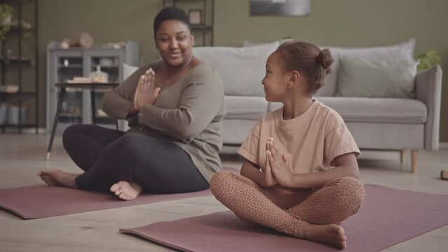 Slowmo of relaxed African-American curvy woman and her 5 year old daughter sitting on yoga mats at home meditating together