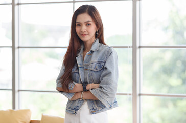 Fototapeta premium Portrait closeup shot of Asian young beautiful long brown hair female teenager fashion model in casual blue street denim jeans jacket standing smiling look at camera holding hand at chin crossed arm