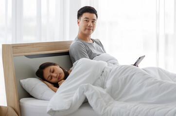 Portrait shot of married couple Asian handsome male husband sitting look at camera on bed watching movie from touchscreen tablet while female wife sleeping sweet dream cover with blanket in bedroom