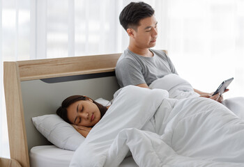 Portrait shot of married couple Asian handsome male husband sitting look at camera on bed watching movie from touchscreen tablet while female wife sleeping sweet dream cover with blanket in bedroom
