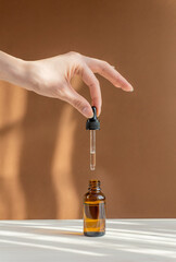 Vertical image of pipette with cosmetic serum in women's hands on light brown background, in rays...