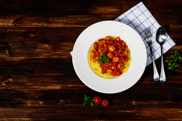 Closeup top view shot of delicious tasty Italian sliced sausages and minced pork meat in red sauce pasta spaghetti on white dish placed on wood table with napkin spoon fork with tomatoes and onions