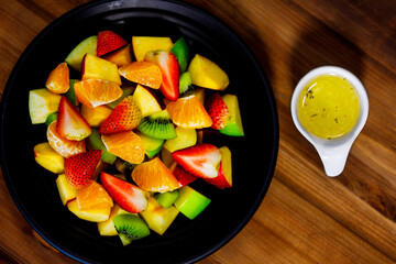 Top view shot of mixed sliced fresh raw healthy tasty fruits salad appetizer and dressing placed on wooden table with spoon fork decorated with sack napkin serving with tomato juice drink in glass