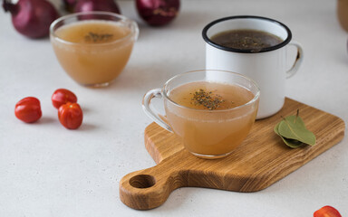 Homemade bone broth in mugs with spices on a wooden stick with bay leaf, tomatoes and onions.