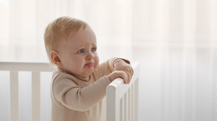 Crying baby portrait. Little infant tired and hungry, start crying standing in crib, looking aside...