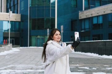 Urban portrait of young elegant business woman in winter casual clothes, white jacket. Walking in the city street, talking on the phone, making selfie. Working outdoor