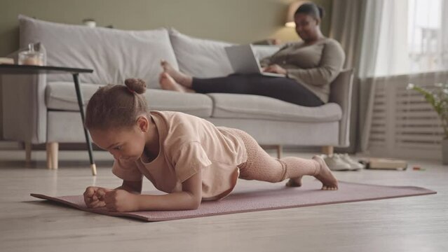 Slowmo of sporty African-American gen A girl doing plank exercise on yoga mat in living room while her mother working on laptop sitting on sofa in background