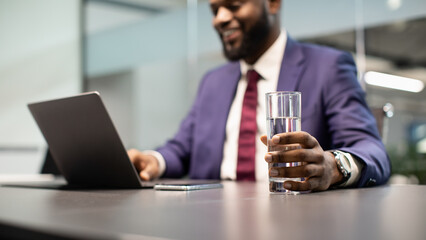 Positive black manager drinking water while working on laptop, panorama