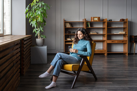 Smiling young woman rest after work in office free space or at home sit in armchair communicating with friends in smartphone app. Girl with mobile phote browsing social media or do online shopping