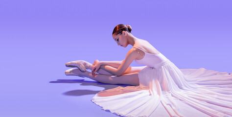 an elegant ballerina in pointe shoes sits stretching her legs in a long white skirt on a lilac...