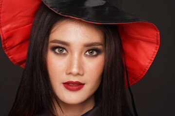 Portrait studio shot of Asian young beautiful makeup female wears witch costume with tall hat and orange rope sitting smiling look at camera on black background in traditional halloween night party