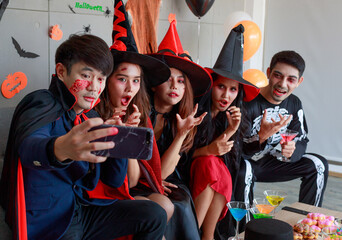 Fototapeta premium Group of Asian young scary creepy male and female friends in Halloween witch sorcerer ghost and skeleton costume sitting on sofa taking selfie photo together in traditional trick or treat night party