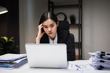 Stressed asian business woman working late at night in the office hands on head feeling headache. Tired woman looking at laptop working hard sitting in the dark room office. Overtime concept