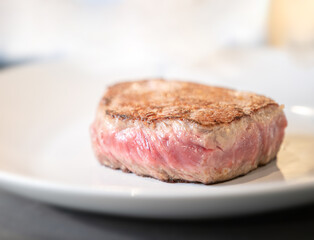 A zoomed-in picture with a blurry background of a medium-cooked beefsteak on a white plate placed on a table.