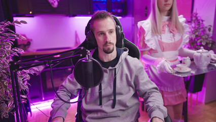 Cute maid in pink uniform serving male gamer tea with kitchen in background