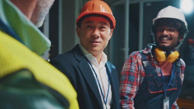Happy funny foreman, asian architect and builder in hardhats and uniforms discussing blueprint. Talking about construction project at construction site in the evening. Slow motion