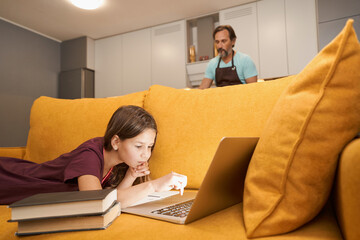 Adult man cooks in the kitchen while his daughter works on a laptop