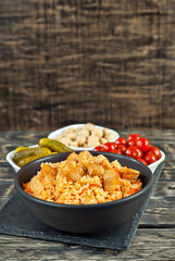 Pilaf with meat and carrots in a black bowl. Marinated mushrooms, tomato and cucumber on a black background. Food on a shabby table. Bowl on an old wooden board.