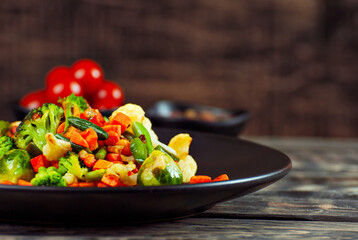 Mix of vegetables close up. Salad of various chopped vegetables. Roasted cabbage, carrots, broccoli, peas in a black plate. Food on a wooden board. Marinated tomatoes in the background.
