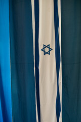 A long, vertical, blue and white flag of the State of Israel on a background of large strips of blue fabric.