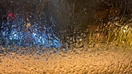 Frozen ice in the bright glow of the light on the car window at night or in the evening out of focus. Drops on glass. Aspect bokeh texture. Winter.