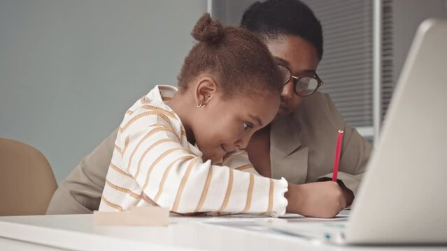 Waist up slowmo of hard-working preschool African-American girl writing on sheet of paper while practicing English alphabet together with female teacher helping her