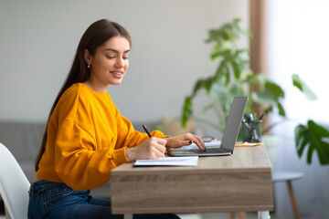 Happy Caucasian woman using laptop pc, working or studying online, taking notes during webinar or remote lesson at home
